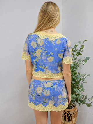 A woman viewed from the back wearing a matching blue floral lace top and skirt set with yellow lace trim, standing indoors next to a potted green plant against a light gray wall.
