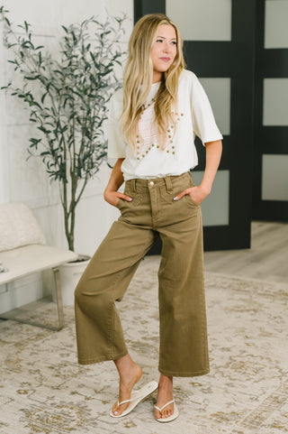 A woman modeling a white short-sleeve t-shirt with gold beaded fringe and wide-leg olive green cropped pants, standing in a modern indoor setting with a patterned rug and a potted plant.