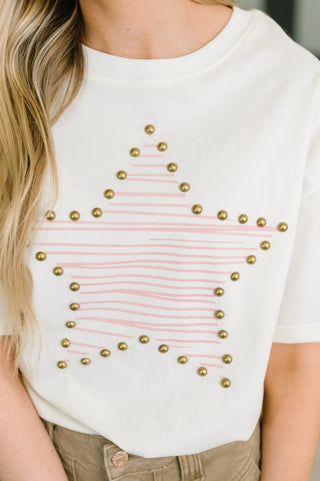 A close-up of a person wearing a white short-sleeve T-shirt with a large outlined star design made of thin pink lines and metallic gold studs, paired with tan pants.