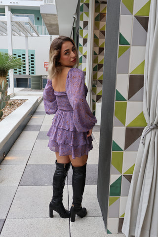 Woman in a purple dress and black knee-high boots standing on a geometric-patterned wall.