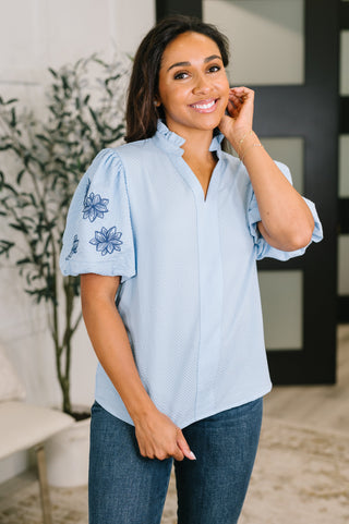 A woman wearing a light blue short-sleeve button-up blouse with blue floral embroidery on the sleeve, paired with dark blue jeans, standing in a modern indoor setting.