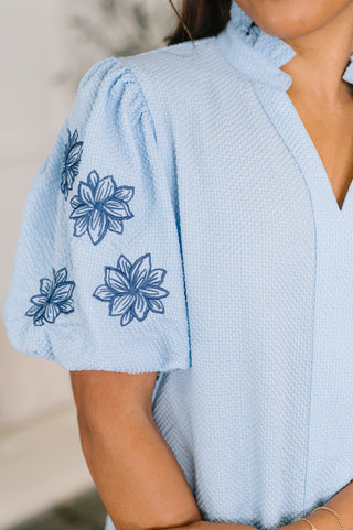 A close-up of a woman wearing a light blue textured blouse with dark blue floral embroidery on the puff sleeves.