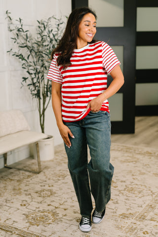 A woman modeling a red and white horizontally striped short-sleeve t-shirt, paired with dark blue jeans and black sneakers, standing in a modern indoor setting with a patterned rug and a potted plant.