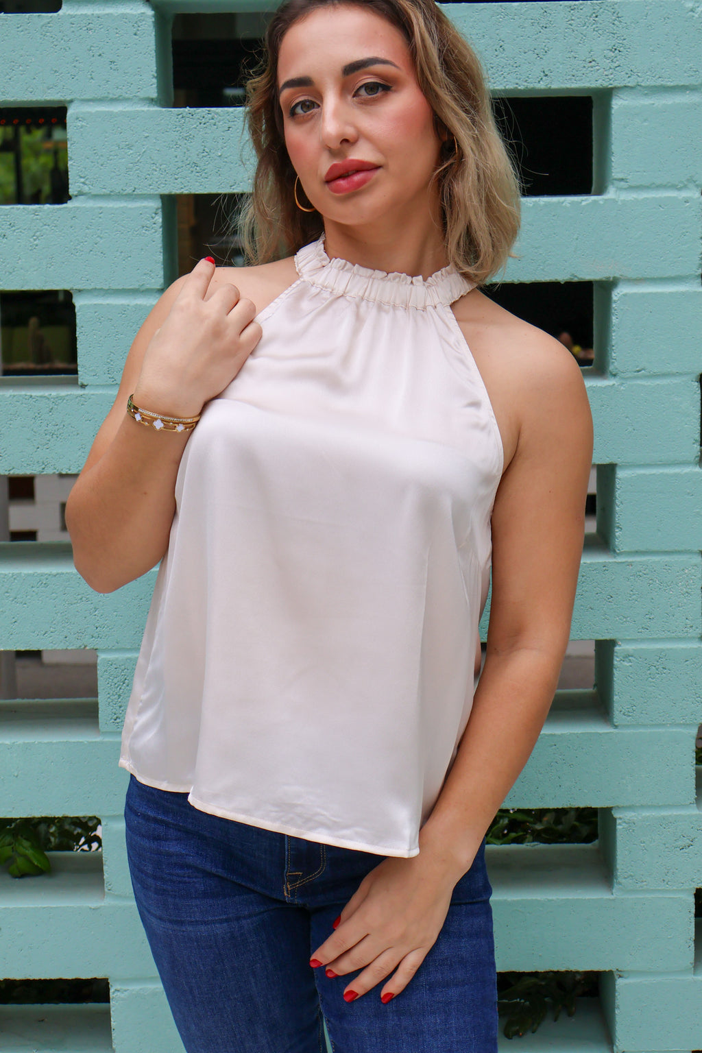 Woman wearing a light beige halter top and blue jeans against a teal brick wall.